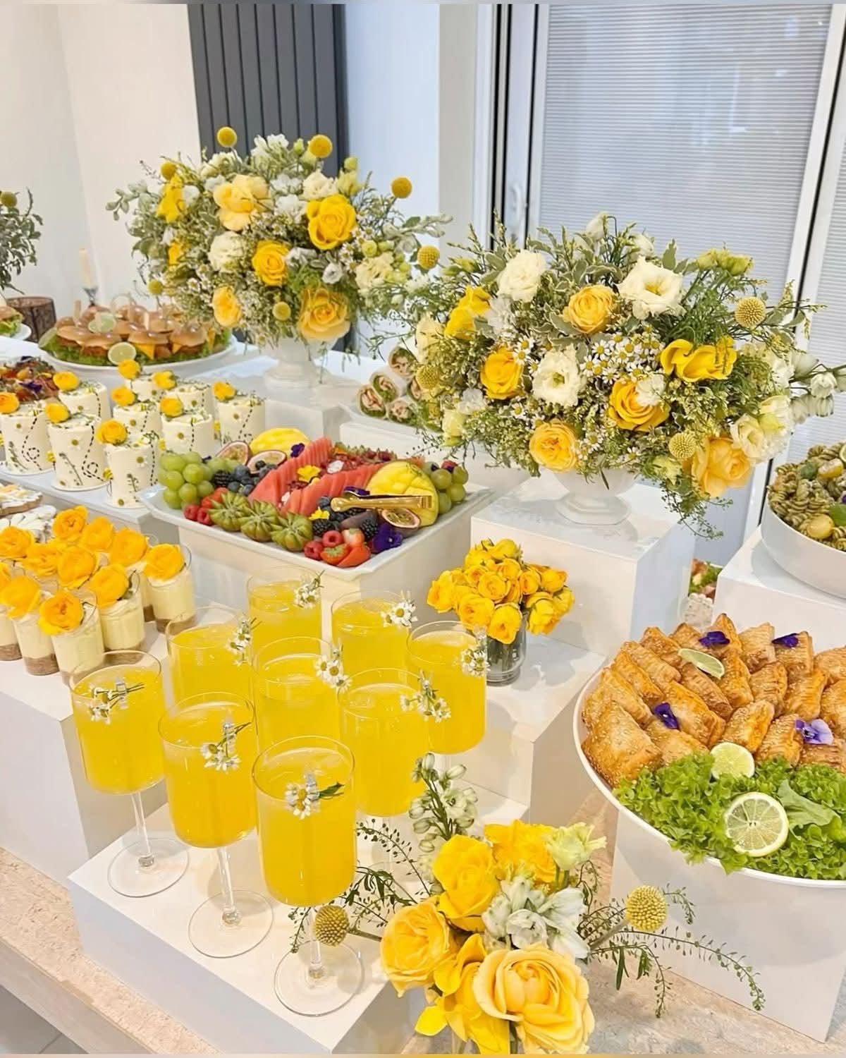 A table decorated with yellow and white floral arrangements, served with assorted fruit, pastries, and yellow drinks, suitable for a celebration or party.