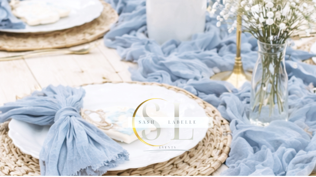 Table setting with a white plate, light blue napkin tied with a knot, a glass vase with white flowers, and a gold candlestick, set on a light wood table with a blue table runner.