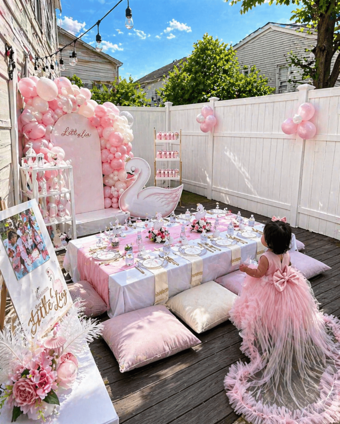 A pink and white themed outdoor birthday party setup with a low table, cushions, and a large swan and balloon decoration, decorated with pink balloons, flowers, and a pink dress for a young girl.
