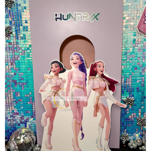 Cardboard cutout of three female characters from the animated series 'HuntX' in front of a colorful tiled backdrop with 'HuntX' logo above.