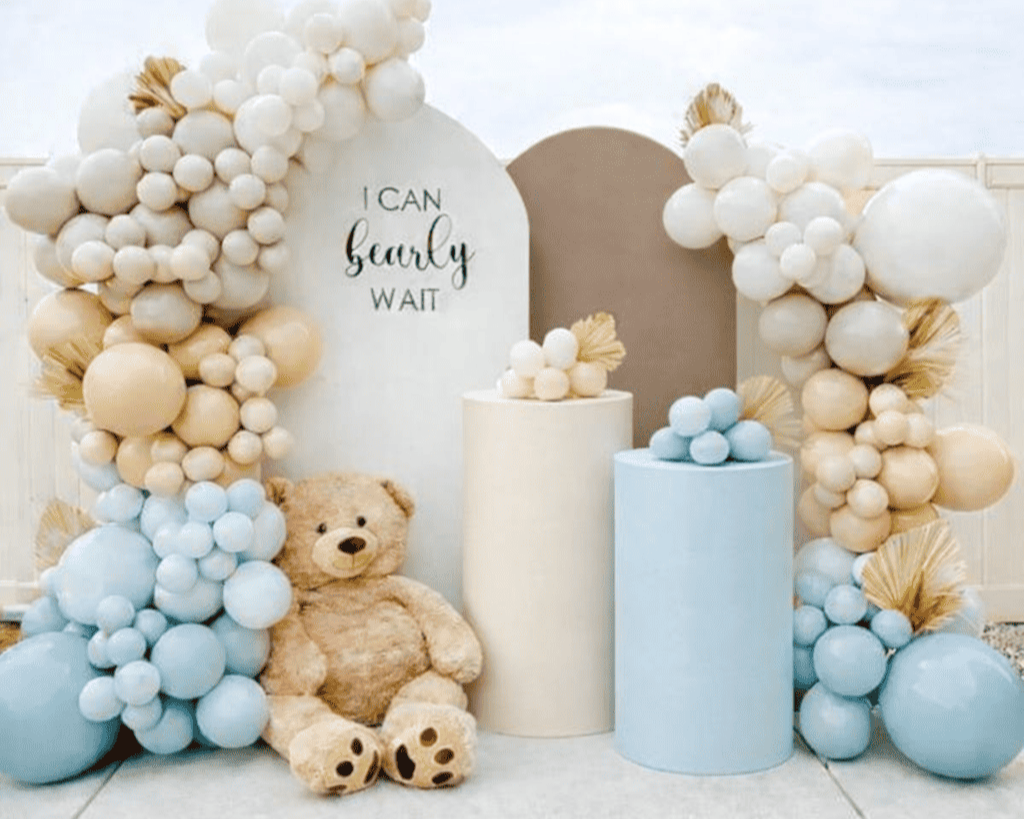 Decorative balloon arch with beige, white, and light blue balloons, a plush teddy bear sitting in front, and a white sign reading 'I CAN hardly WAIT' in elegant script, set against a neutral backdrop.