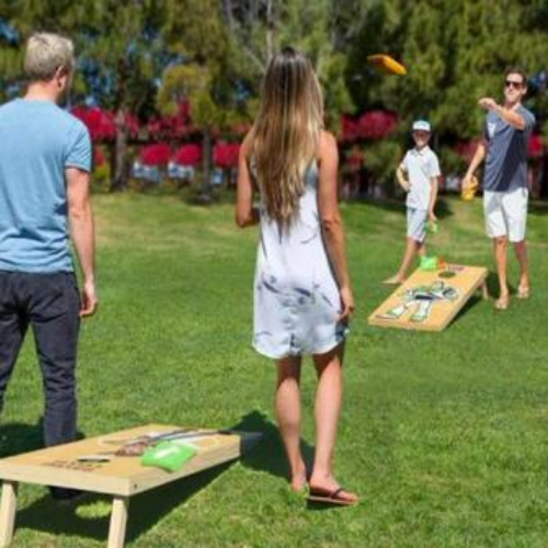 Four people playing cornhole outdoors on a grassy lawn with trees and bushes in the background. A woman in a white dress throws a bean bag, while a man in a gray shirt and white shorts prepares to throw, and another man in a blue shirt stands nearby.
