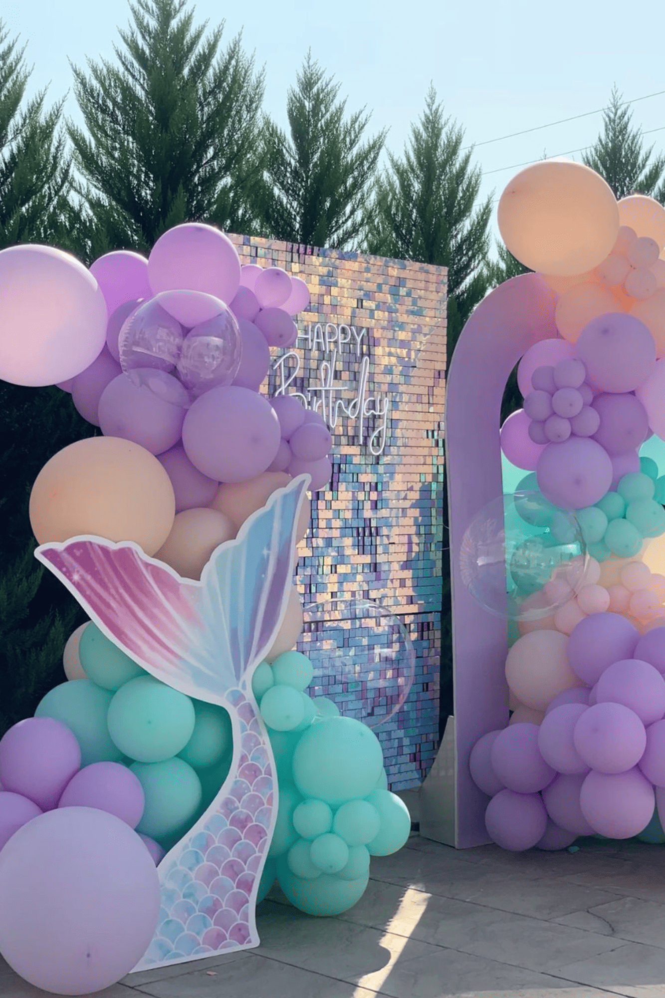 Decorative pastel-colored balloons and a sign that reads 'Happy Birthday' against a brick wall and trees.
