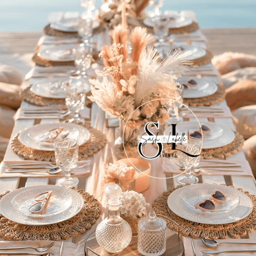 Elegant outdoor dining table set with decorative place settings,1 with heart-shaped chocolate cookies and sunglasses resting on top, surrounded by glassware and adorned with a floral centerpiece of pampas grass and white flowers.