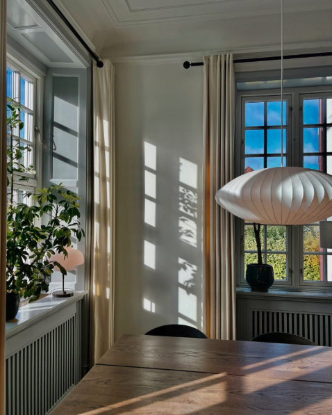 Indendørs spiseområde med vinduer, gardiner og et rundt loftlys.