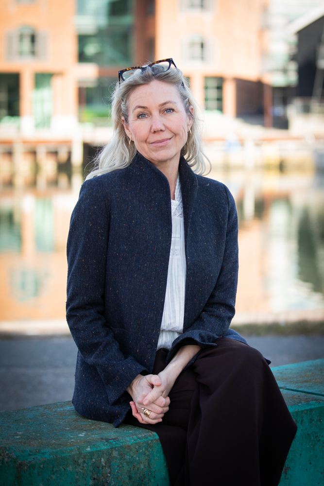 Karen Schou iført en mørk blazer og hvide skjorte, baggrund med moderne boligbyggeri og vand.