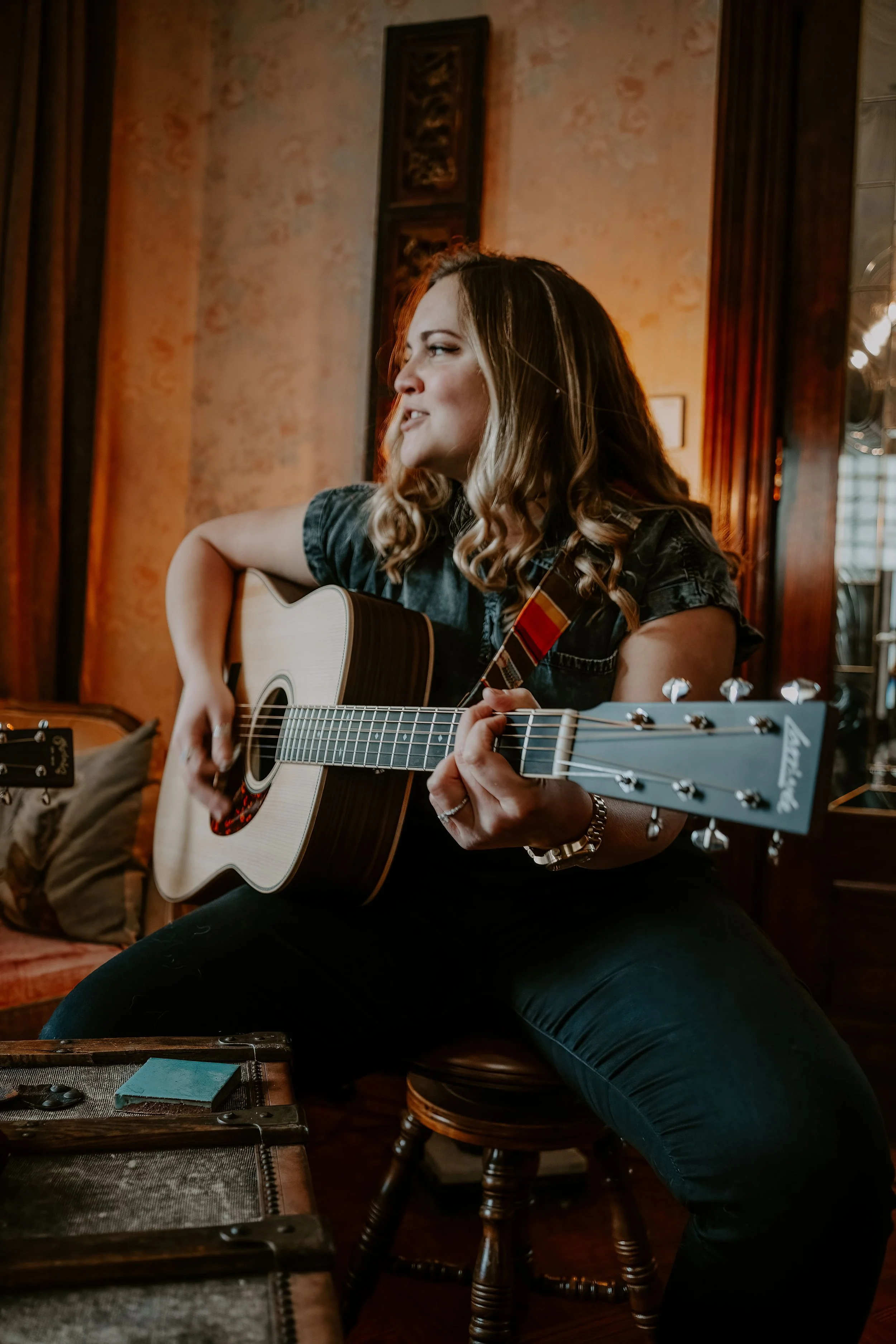 A woman with long, wavy blonde hair playing an acoustic guitar in a cozy, warmly lit room with wooden paneling and framed artwork on the walls.