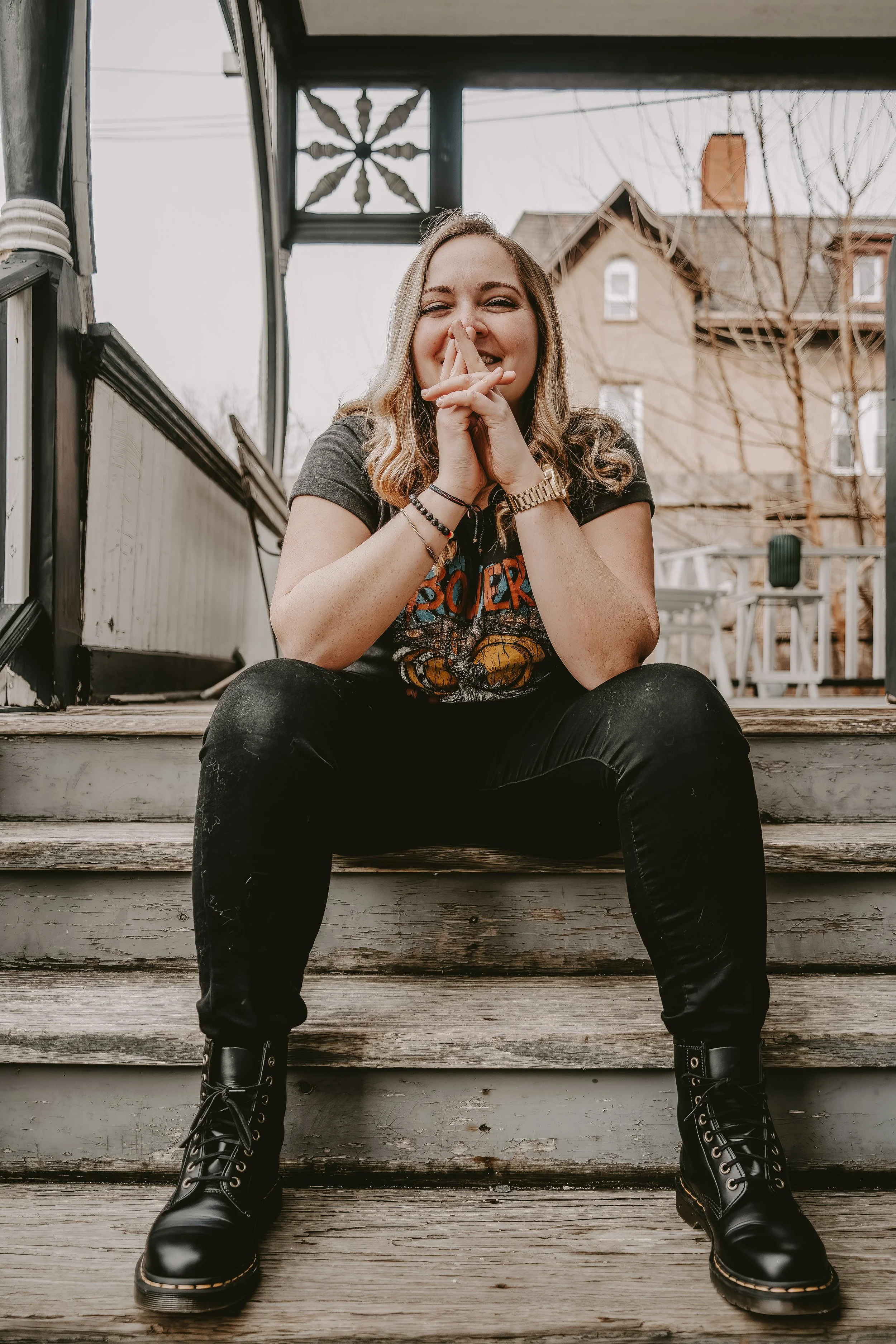 Young woman sitting on wooden steps outdoors, laughing and covering her mouth with her hands, wearing a graphic T-shirt, black pants, and combat boots, with a house and leafless trees in the background.