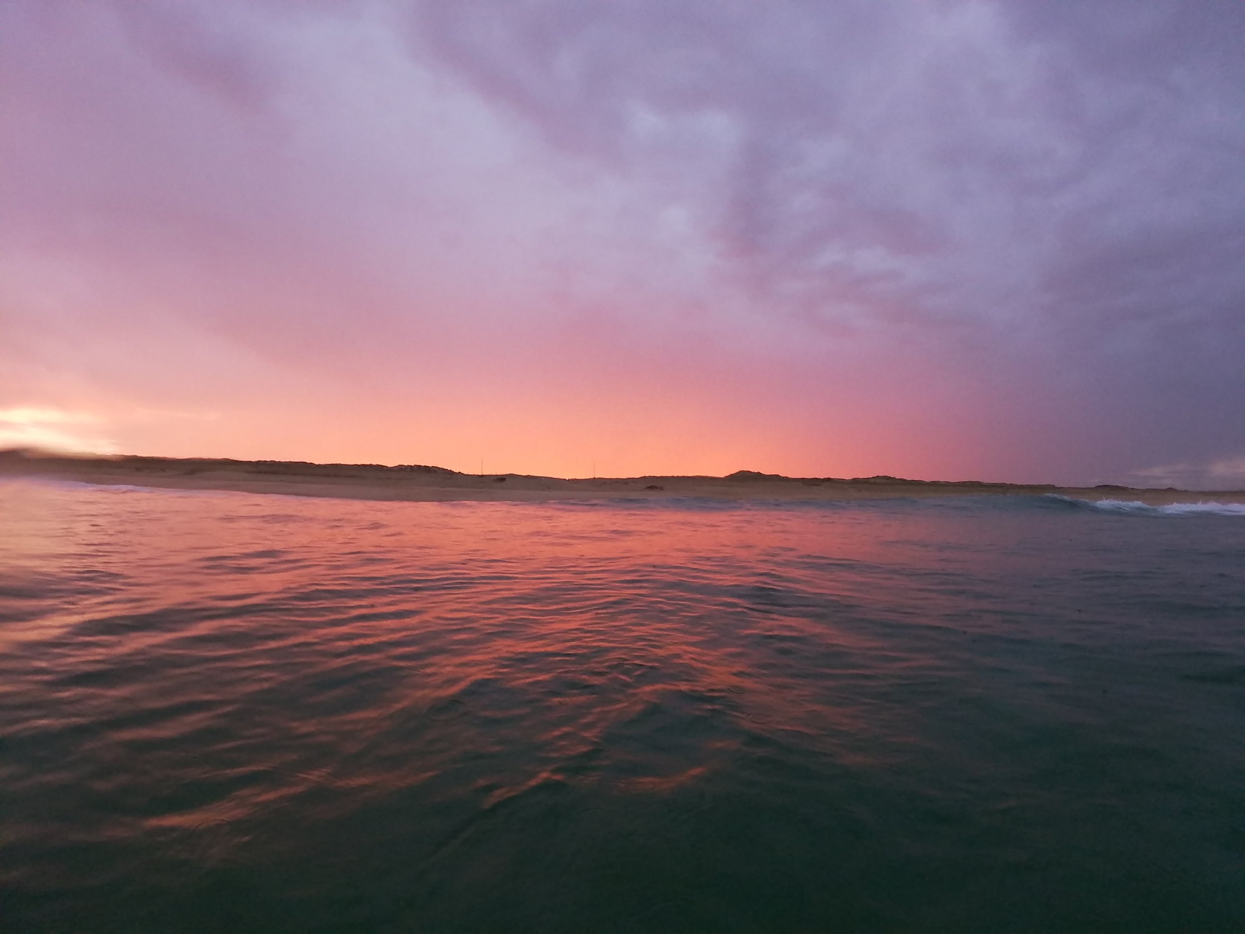 Coucher de soleil sur l'océan avec des nuages violets et roses, horizon lointain avec dunes ou terres pfombres.