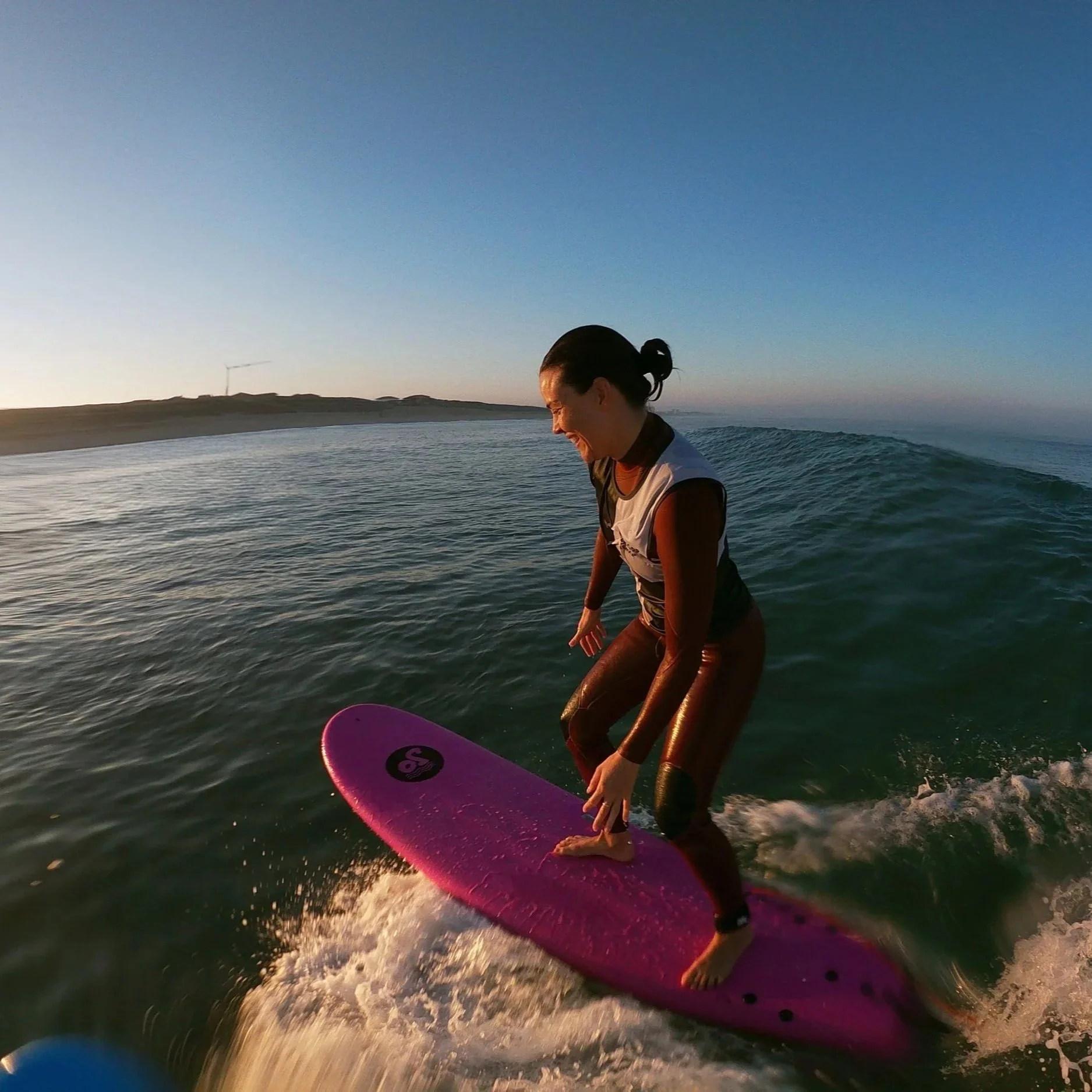 Une personne pratique le surf sur une planche rose dans l'océan, au coucher du soleil, avec un sourire et un paysage côtier en arrière-plan.