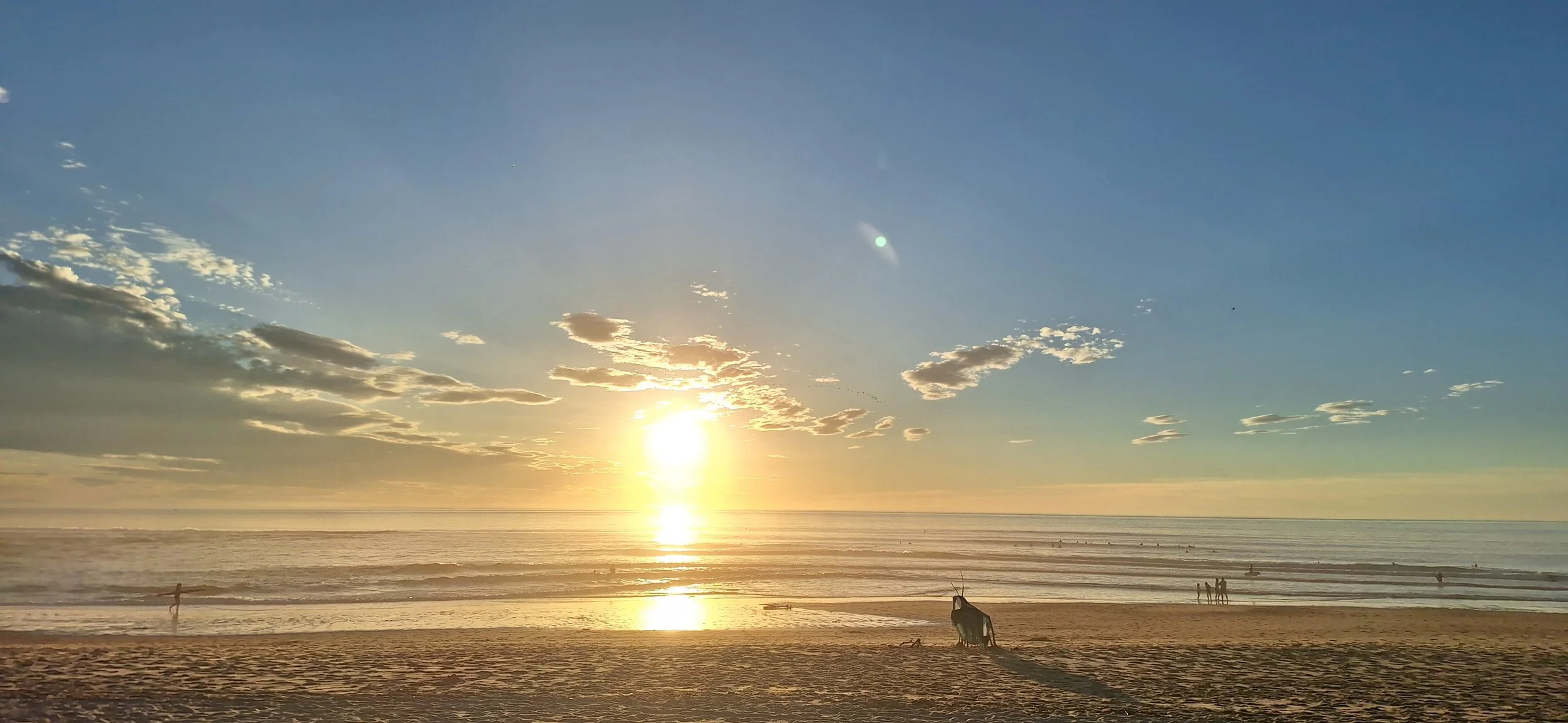 Coucher de soleil sur la plage avec quelques nuages dans le ciel, des silhouettes de personnes et de planches de surf sur le sable.
