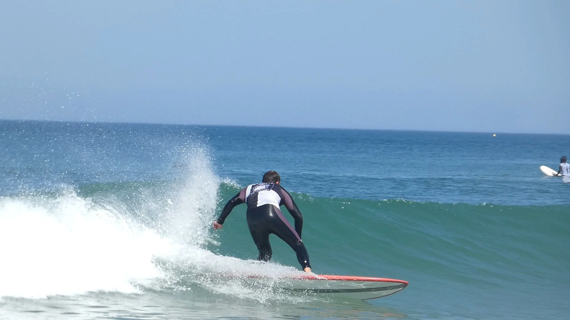 Un surfeur en wetsuit navigue sur une vague dans l'océan, avec deux autres surfeurs au loin, dont un à droite avec une planche blanche.