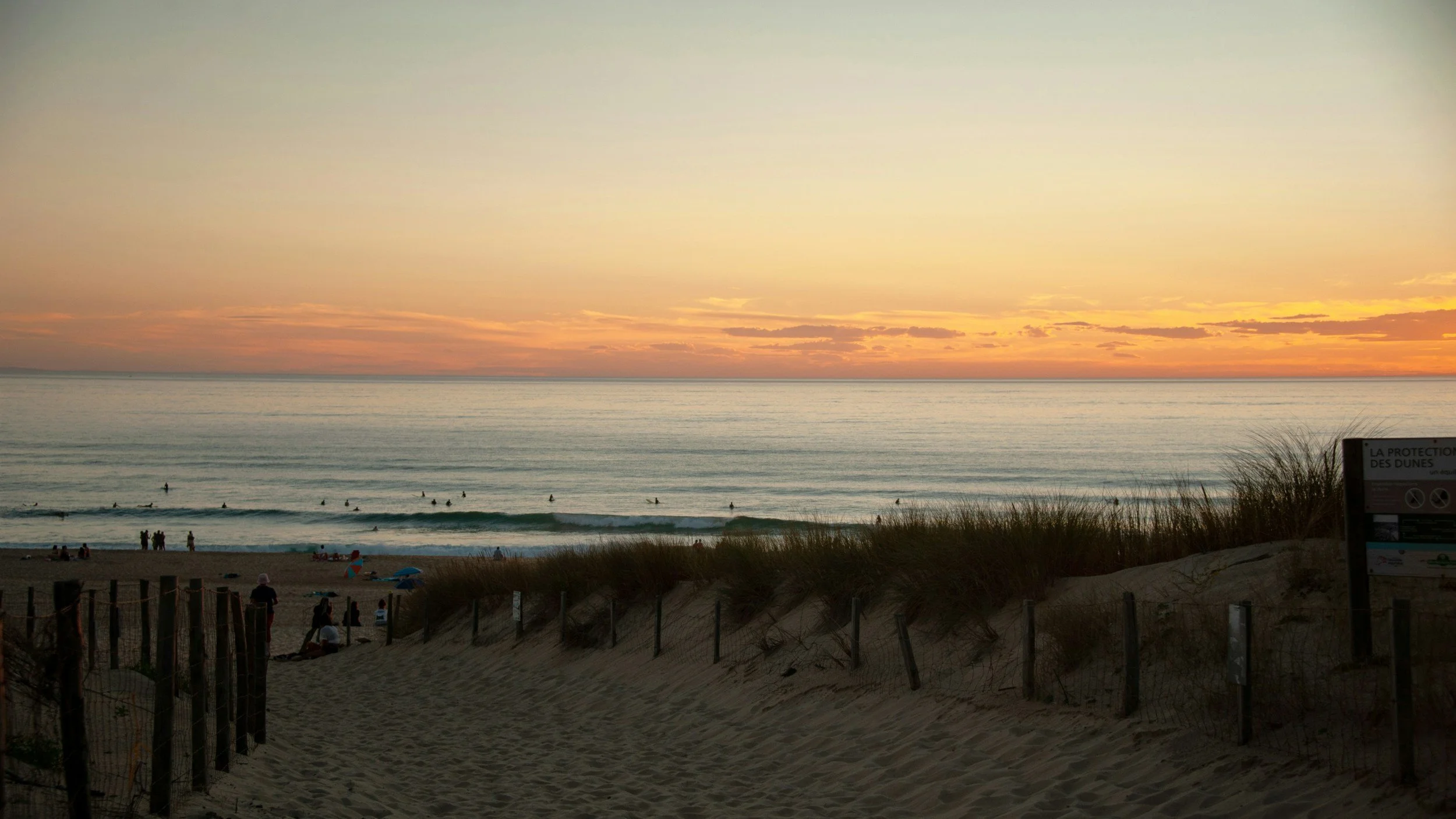 Coucher de soleil sur la mer avec des vagues, des personnes sur la plage, et un panneau de protection des dunes en arrière-plan.