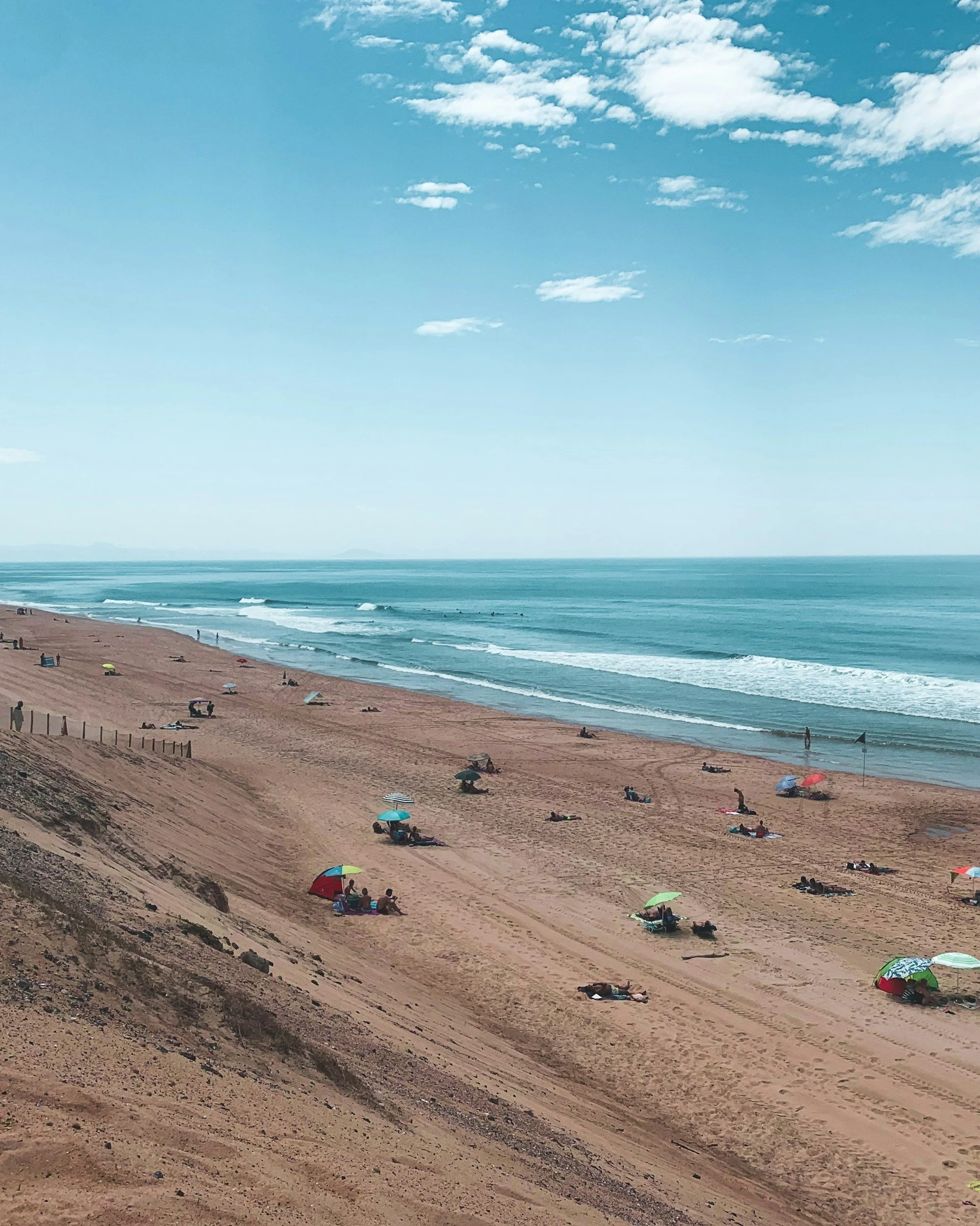 Plage du Santocha à Capbreton avec des parasols colorés et des gens qui se baignent dans l'océan, ciel bleu avec quelques nuages.