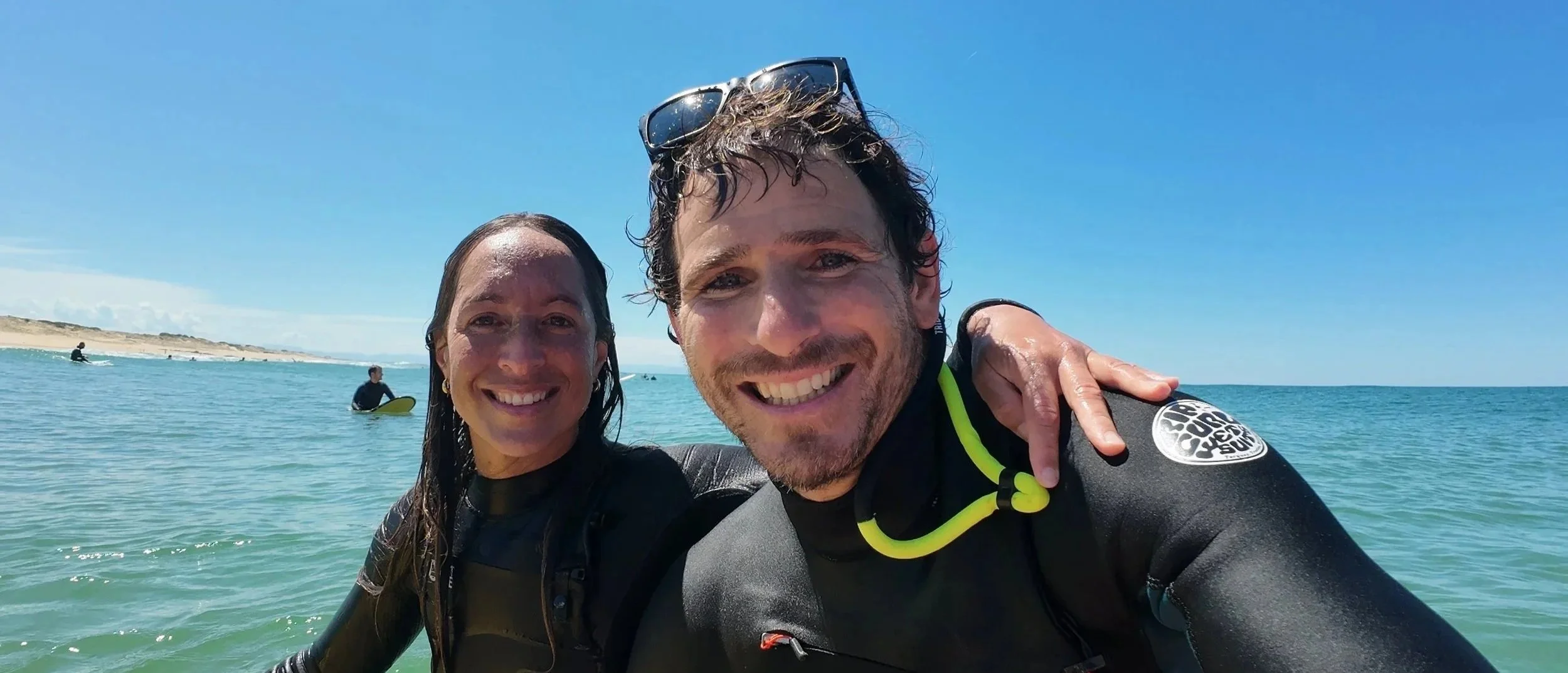 Deux personnes en combinaison de surf souriantes posent pour une photo sur la plage, avec la mer et d'autres surfeurs en arrière-plan, sous un ciel clair.