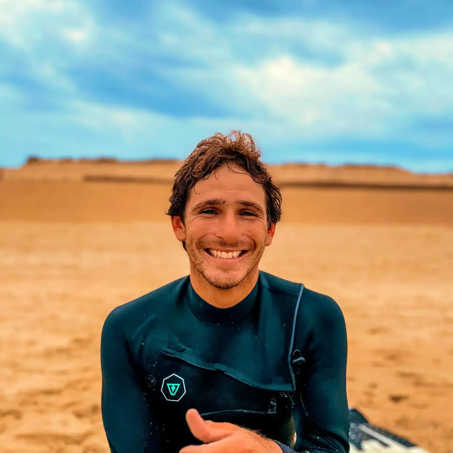 La professeur de surf de l'école 'Hossegor Surf Rendez-vous' portant une combinaison de surf dans une plage désertique avec un ciel bleu et des dunes en arrière-plan.
