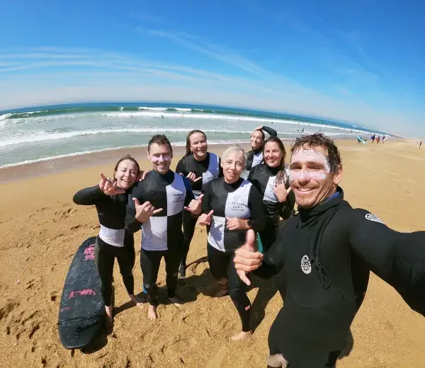 Groupe de six personnes en wetsuit sur la plage, prenant une photo de groupe devant l'océan avec des vagues et un ciel bleu.
