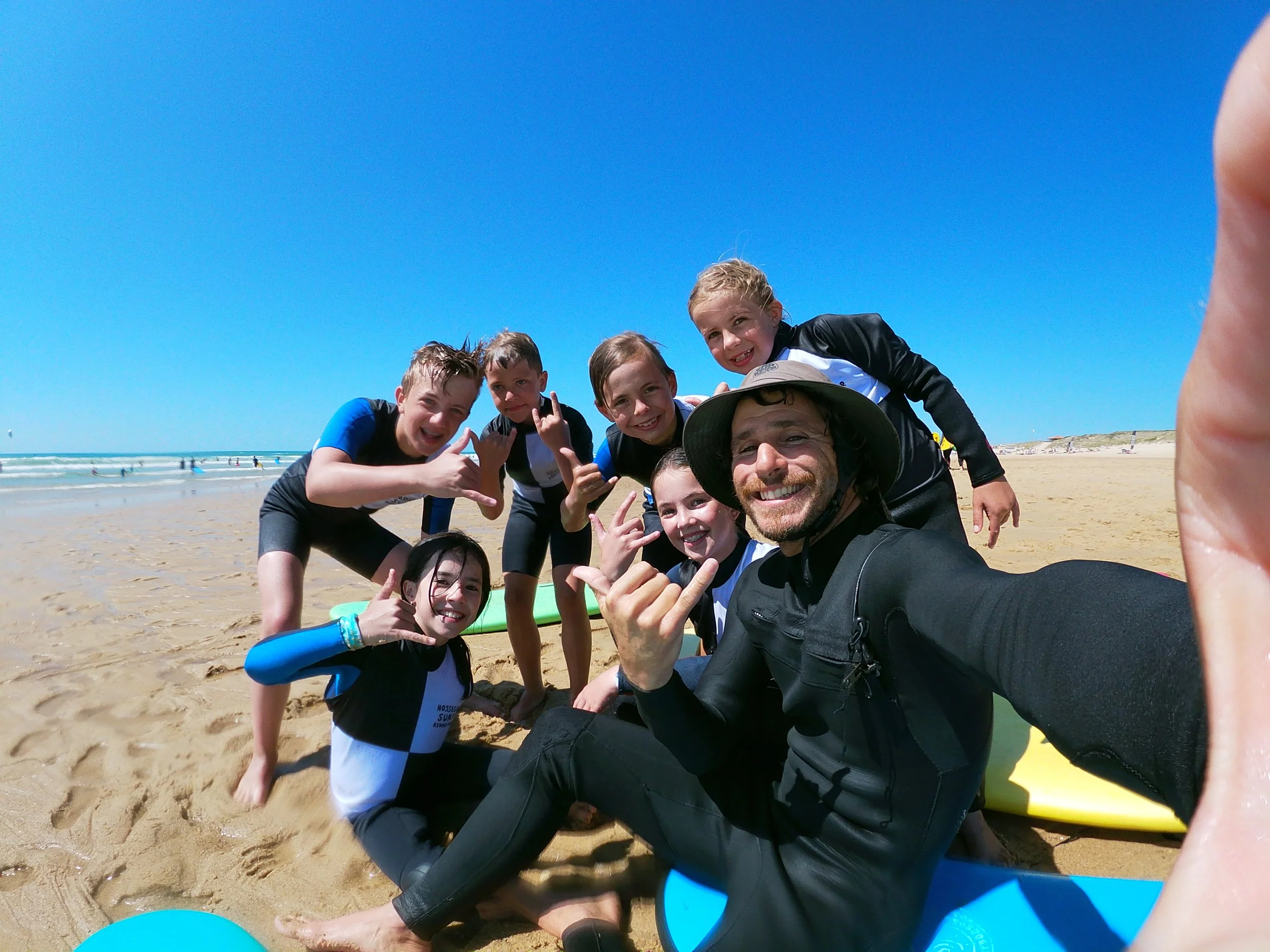 Groupe d'enfants après un cours de surf et le moniteur de surf posant sur la plage sous un ciel bleu, certains faisant des gestes de bras, tous souriants.