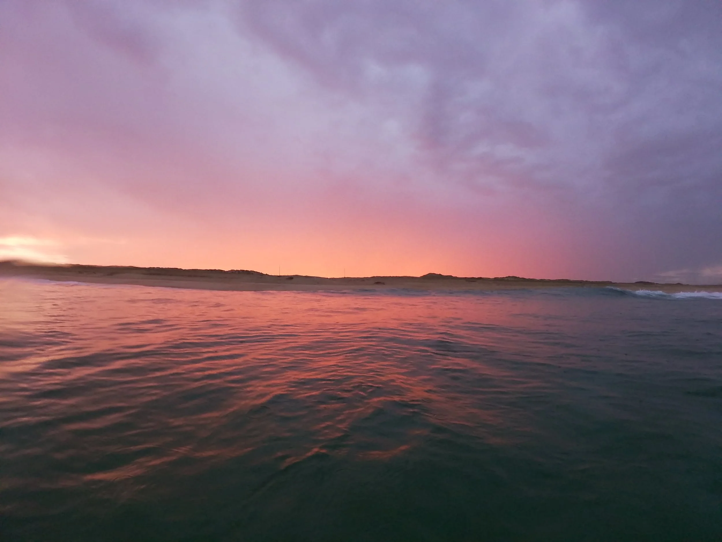 Coucher de soleil sur la mer avec des eaux calmes et un ciel nuageux teinté de couleurs pastel, notamment rose, violet et orange.