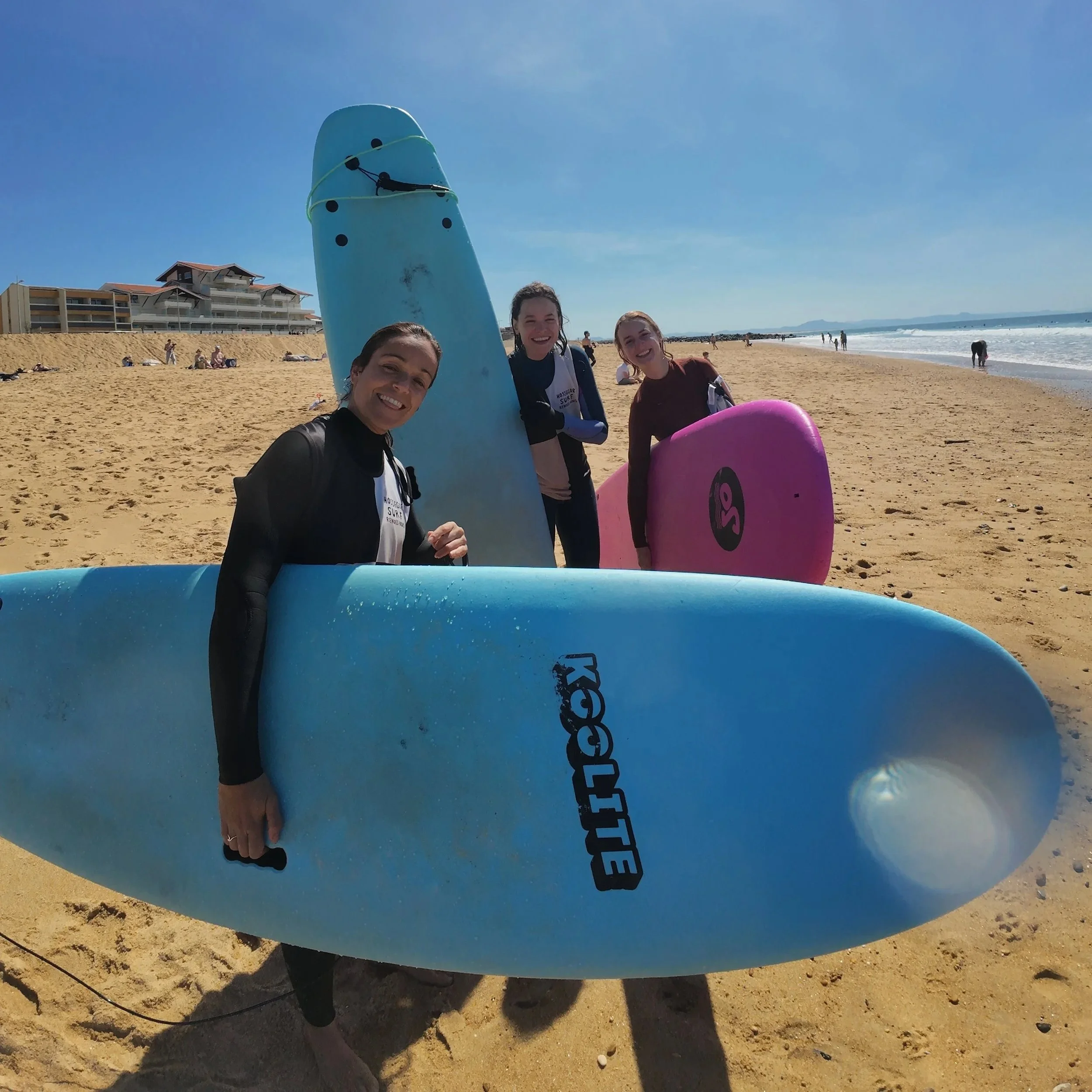 Groupe de trois jeunes femmes souriantes avec des planches de surf sur la plage de sable, ciel bleu clair et bâtiment résidentiel en arrière-plan.