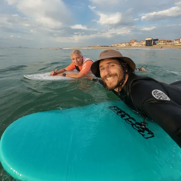 Deux personnes en maillot de bain et équipement de surf, profitant d'une session de surf en mer avec un fond de plage et de bâtiments, sous un ciel partiellement nuageux.