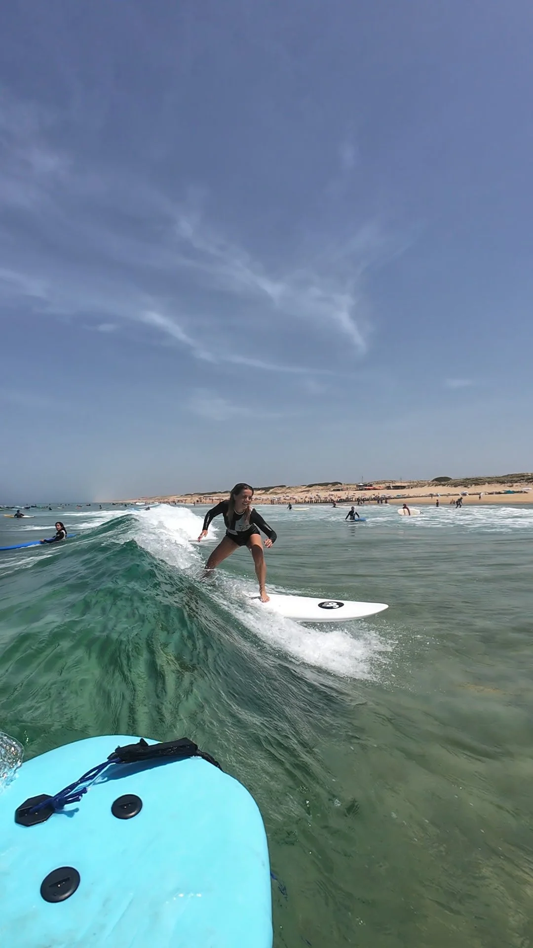 Une femme glisse sur une planche de surf dans la mer, entourée d'autres surfeurs et spectateurs sur la plage, ciel bleu.