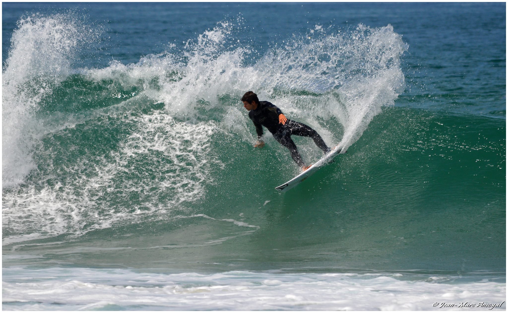 Un surfeur en combinaison noire sur une planche à roulettes, chevauche une vague dans l'océan avec une éclaboussure d'eau derrière lui.