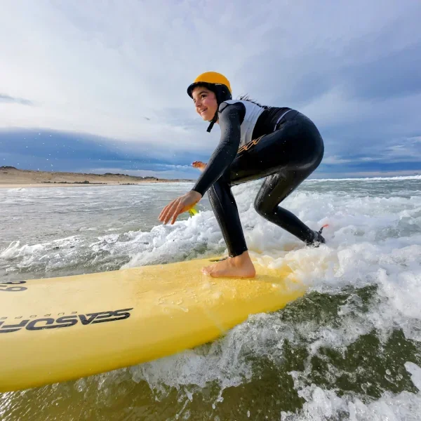 Jeune femme en combinaison de surf et casque de protection sur une planche à surf jaune, surfant sur une vague