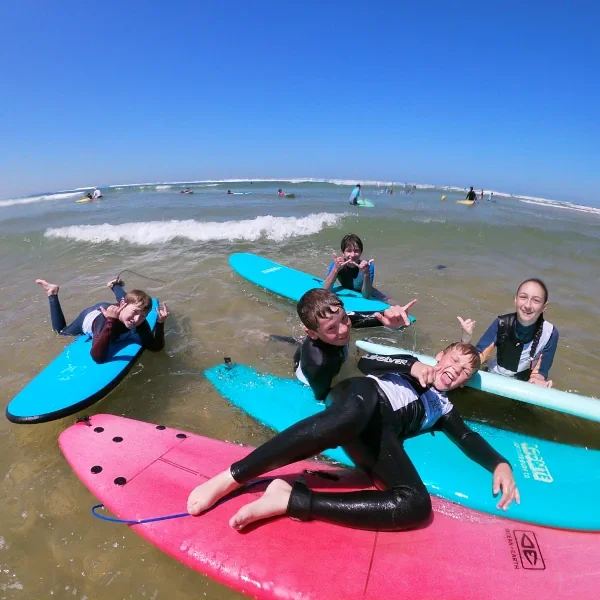 Enfants en train de faire du surf sur la plage, se détendant dans l'eau avec des planches de surf colorées, sous un ciel bleu ensoleillé.
