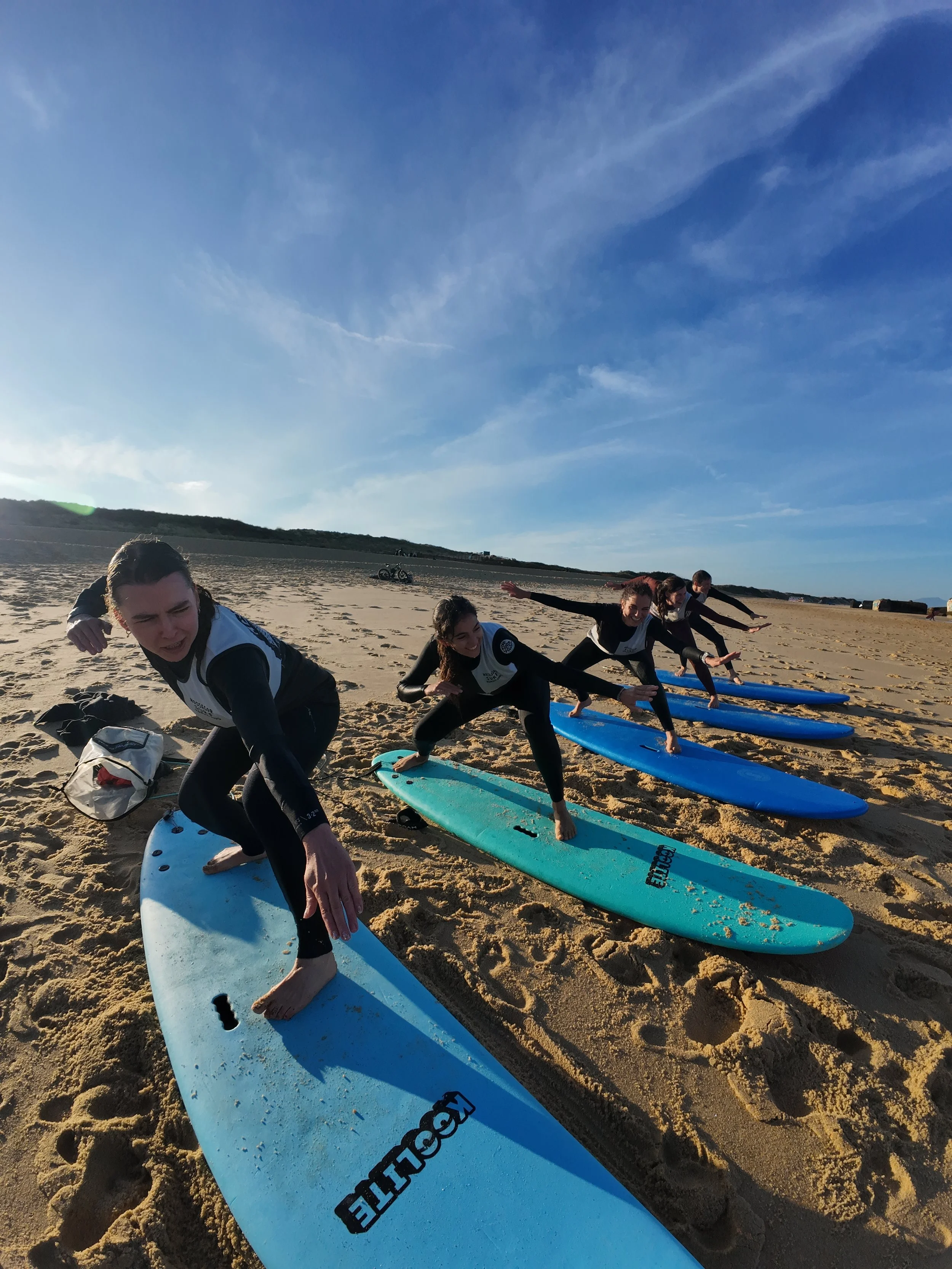 Groupe de personnes faisant du surf debout sur la plage, préparant ou finissant une session de surf avec des planches sur le sable. Le ciel clair et le paysage marin sont visibles en arrière-plan.