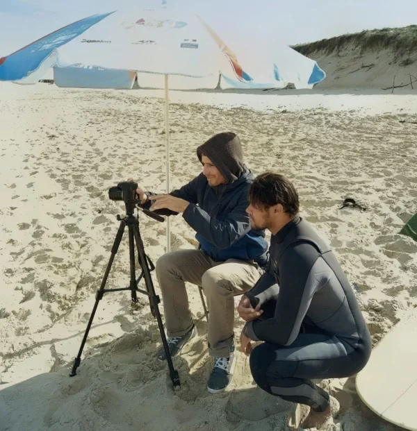 Deux jeunes hommes sur la plage, l'un ajustant un appareil photo sur un trépied, sous un parasol, avec un bateau renversé en arrière-plan.