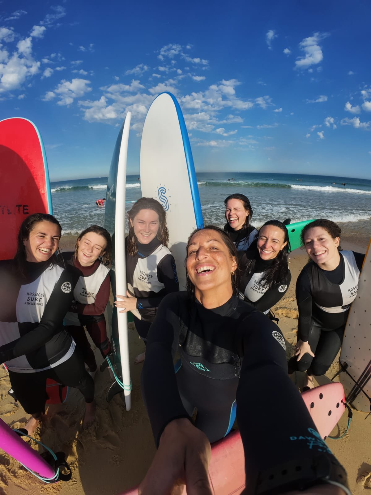 Groupe de femmes souriantes en combinaison de surf avec planches de surf sur la plage, ambiance ensoleillée et ciel bleu.