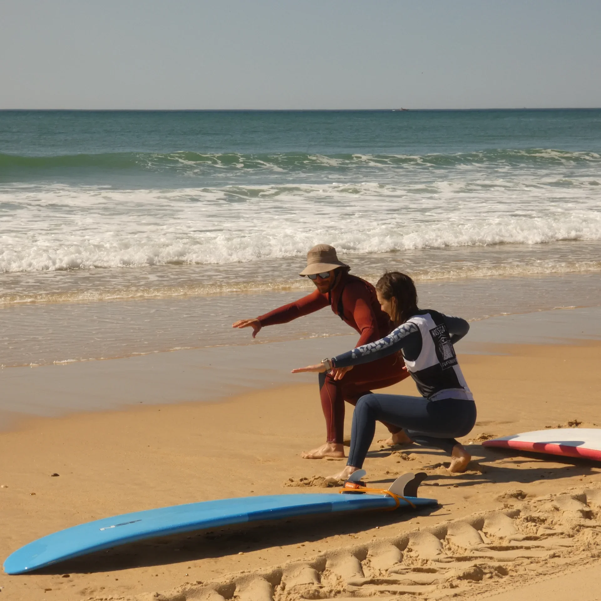 Deux personnes près de la plage, une de face et l'autre de dos, préparant des planches de surf sur le sable, avec la mer en arrière-plan.