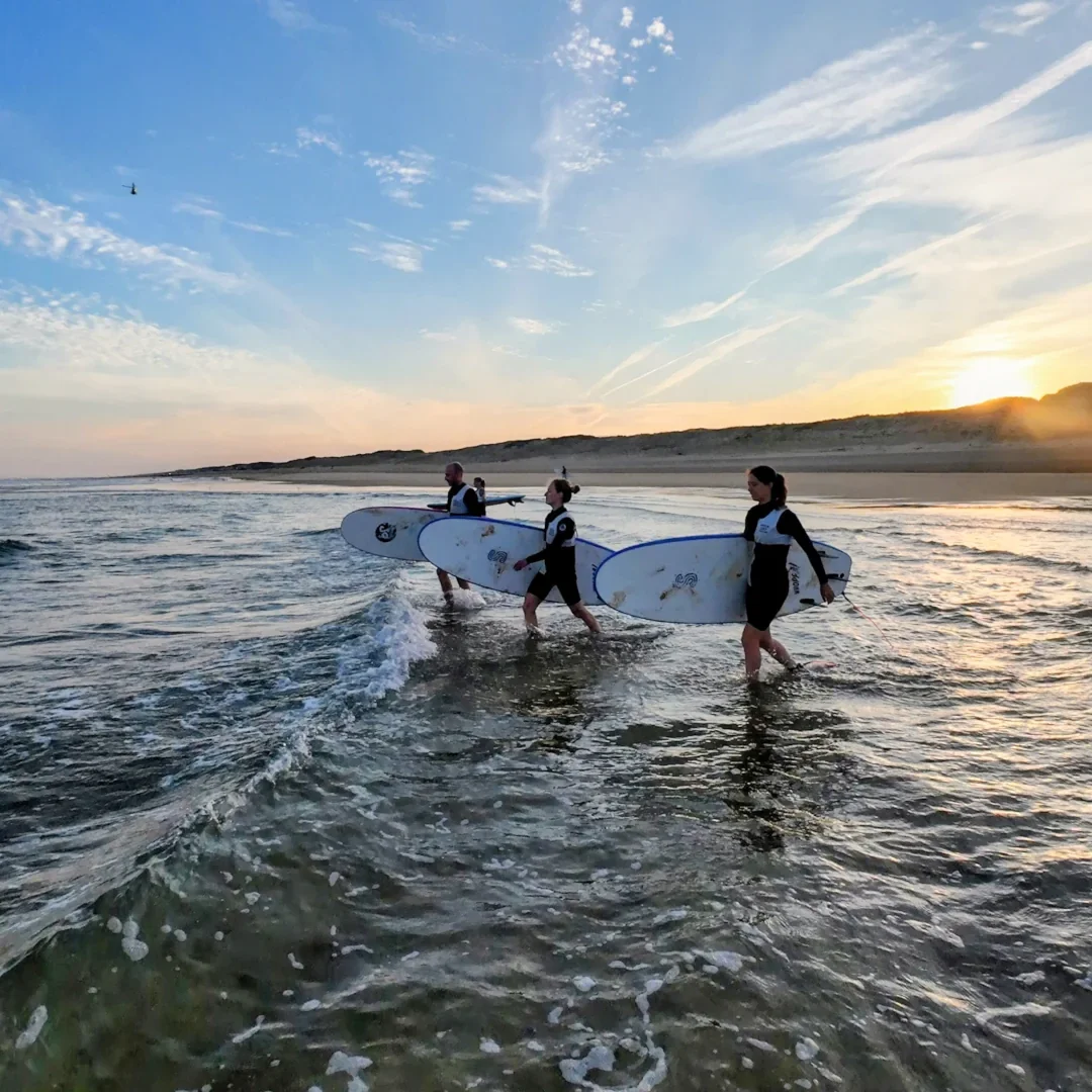Trois personnes avec des planches de surf marchent dans l'eau au coucher du soleil à la plage.