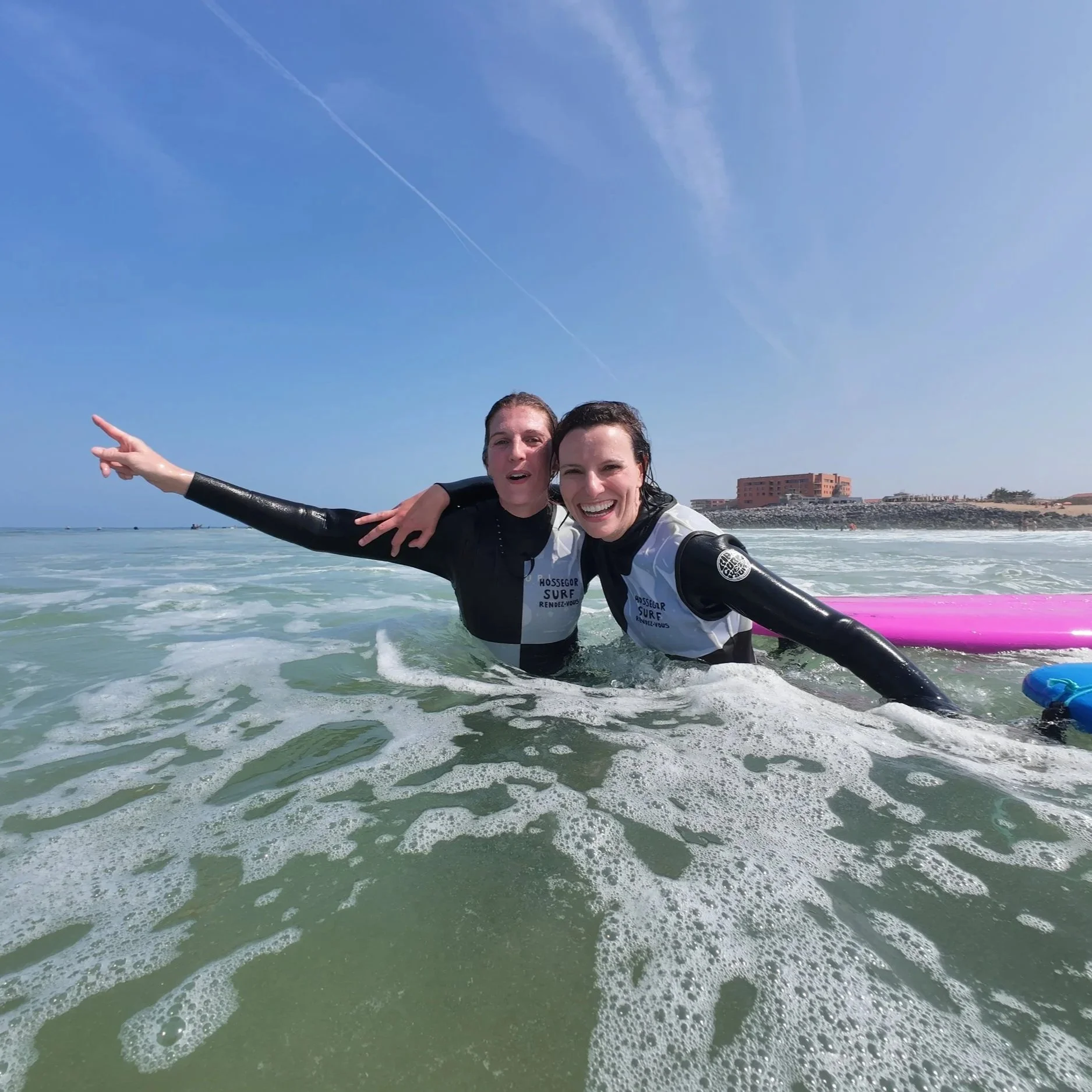 Deux femmes souriantes dans l'eau de la mer, portant des combinaisons de plongée, en train de faire du surf ou de la plongée, avec un ciel bleu clair et un bâtiment en arrière-plan.