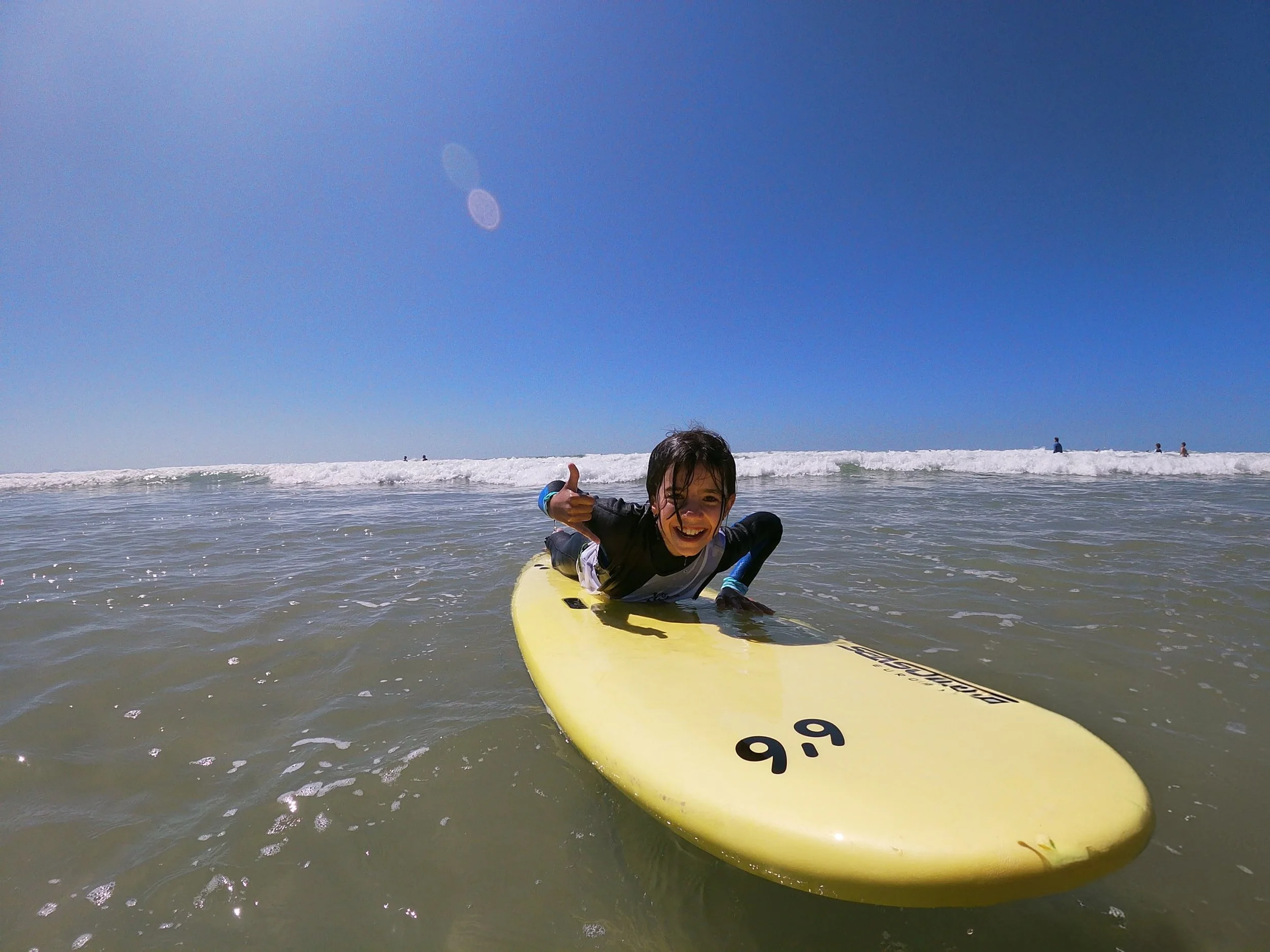 Enfant souriant sur une planche de surf à la plage, donnant un pouce en l'air par temps ensoleillé avec un ciel bleu et des vagues en arrière-plan.