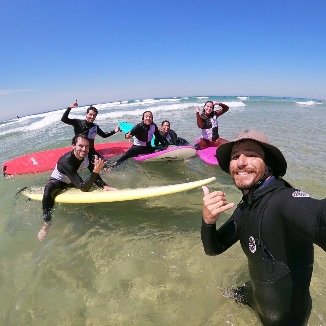 Groupe de personnes en combinaison de surf, tenant des planches de surf, dans l'océan, souriants et faisant des gestes amicaux, avec un ciel bleu clair au-dessus.
