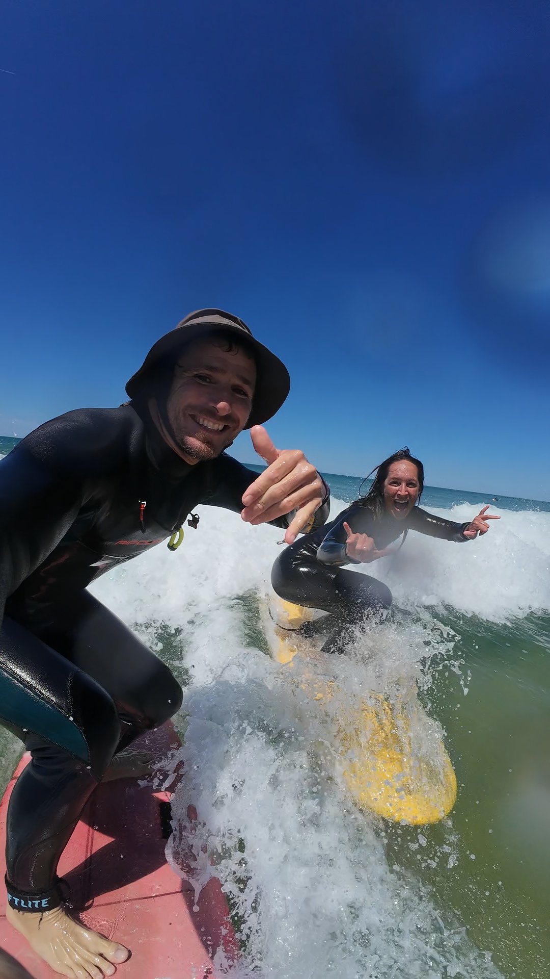 Deux personnes en combinaison de surf sur une planche de surf, souriantes et faisant des gestes de paix, prennent une photo sur une plage en compagnie des vagues de l'océan.