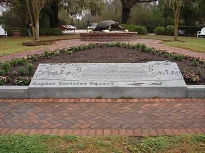 A memorial stone dedicated to the Gopher Tortoise Squaro, surrounded by a flower bed and brick pathway in a park setting.