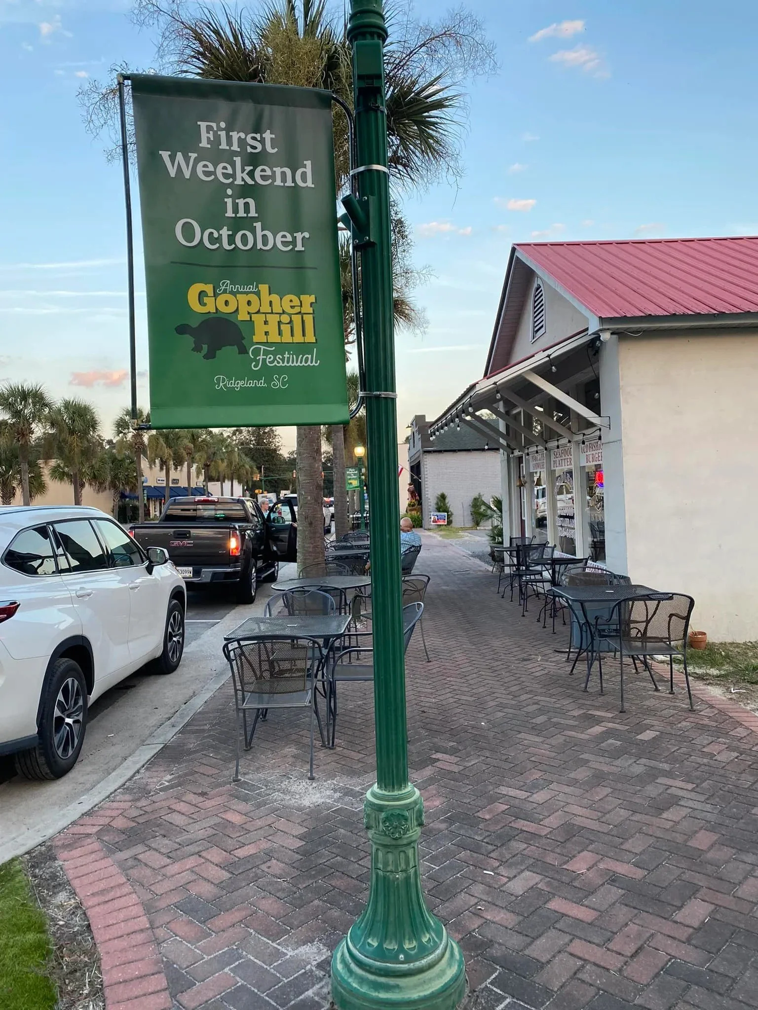 A green lamppost with a banner advertising the first weekend in October for the Annual Gopher Hill Festival in Ridgeland, South Carolina. The background shows parked cars, a building with outdoor seating, and some trees.