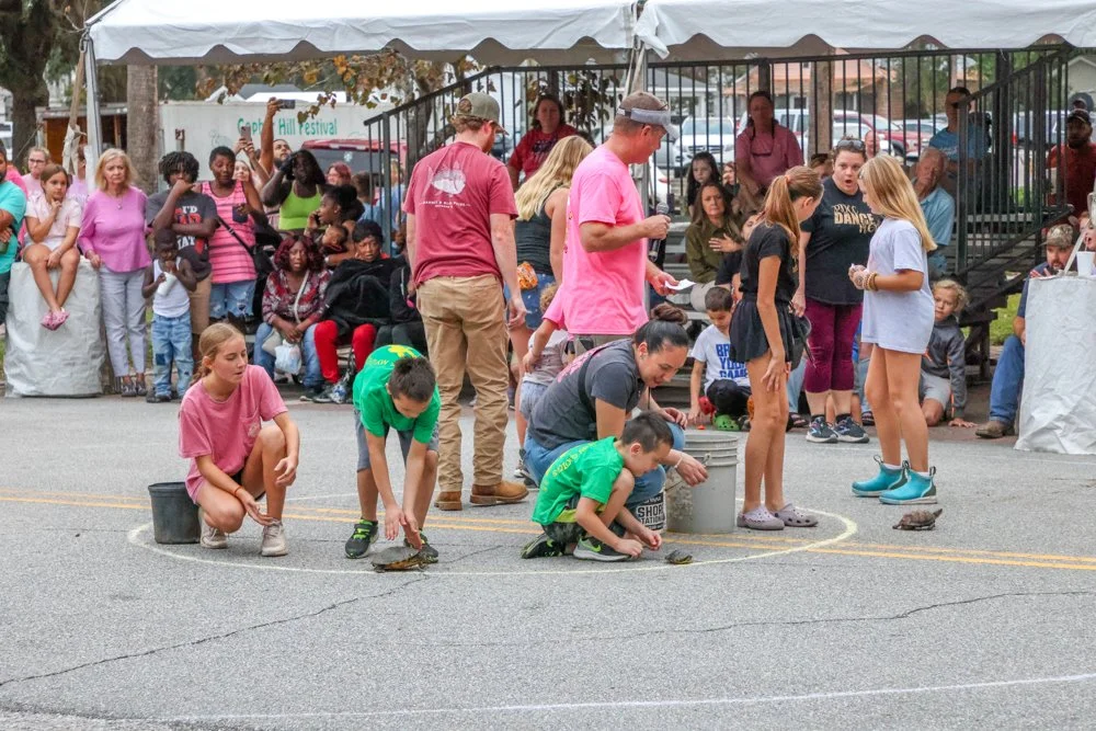 Children and adults participating in a turtle race at a festival, with spectators watching from behind a tent and a railing.