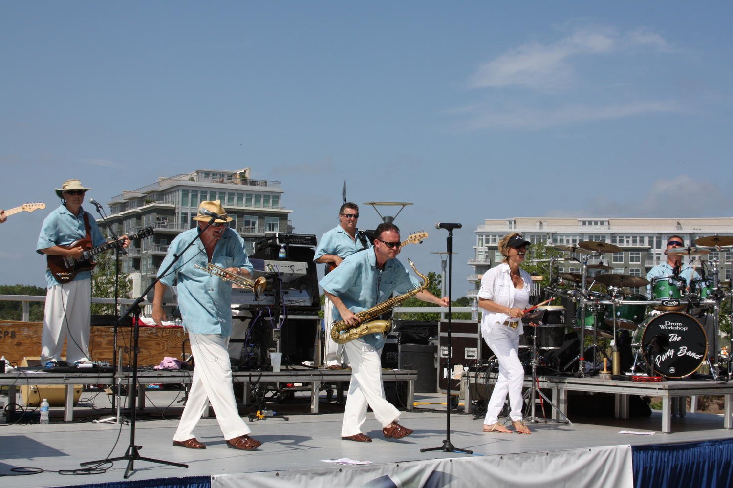 A band performing live on an outdoor stage with musical instruments including guitars, saxophone, drums, and keyboard, under a partly cloudy sky with modern buildings in the background.