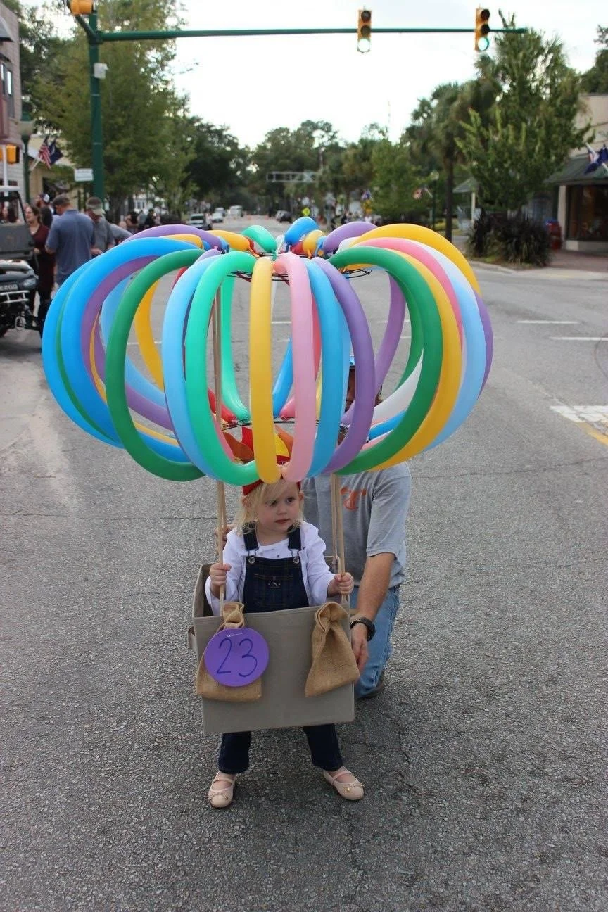 A young girl in a stroller with a cardboard sign labeled '23' is walking in the street during a parade, with a large bunch of colorful balloon animals floating above her, and a man kneeling behind her assisting.
