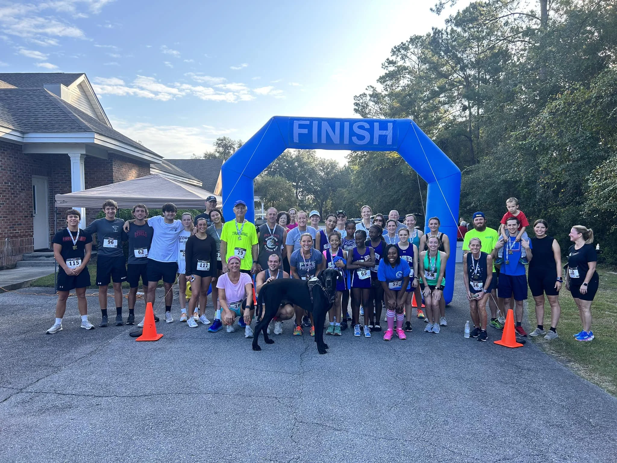 Group of people after a race, standing under a blue finish line arch in front of a brick house and trees, some wearing athletic clothes and medals, with kids and a dog.