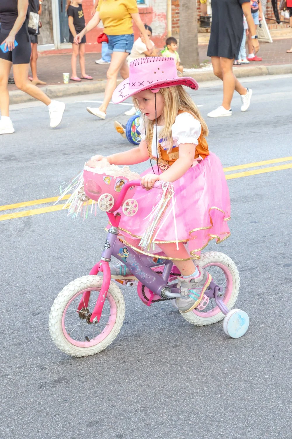 A young girl riding a pink and purple bicycle with training wheels on a street during a parade or event. She is dressed in a princess costume with a pink skirt and a pink cowboy hat. There are other people walking in the background on the sidewalk.