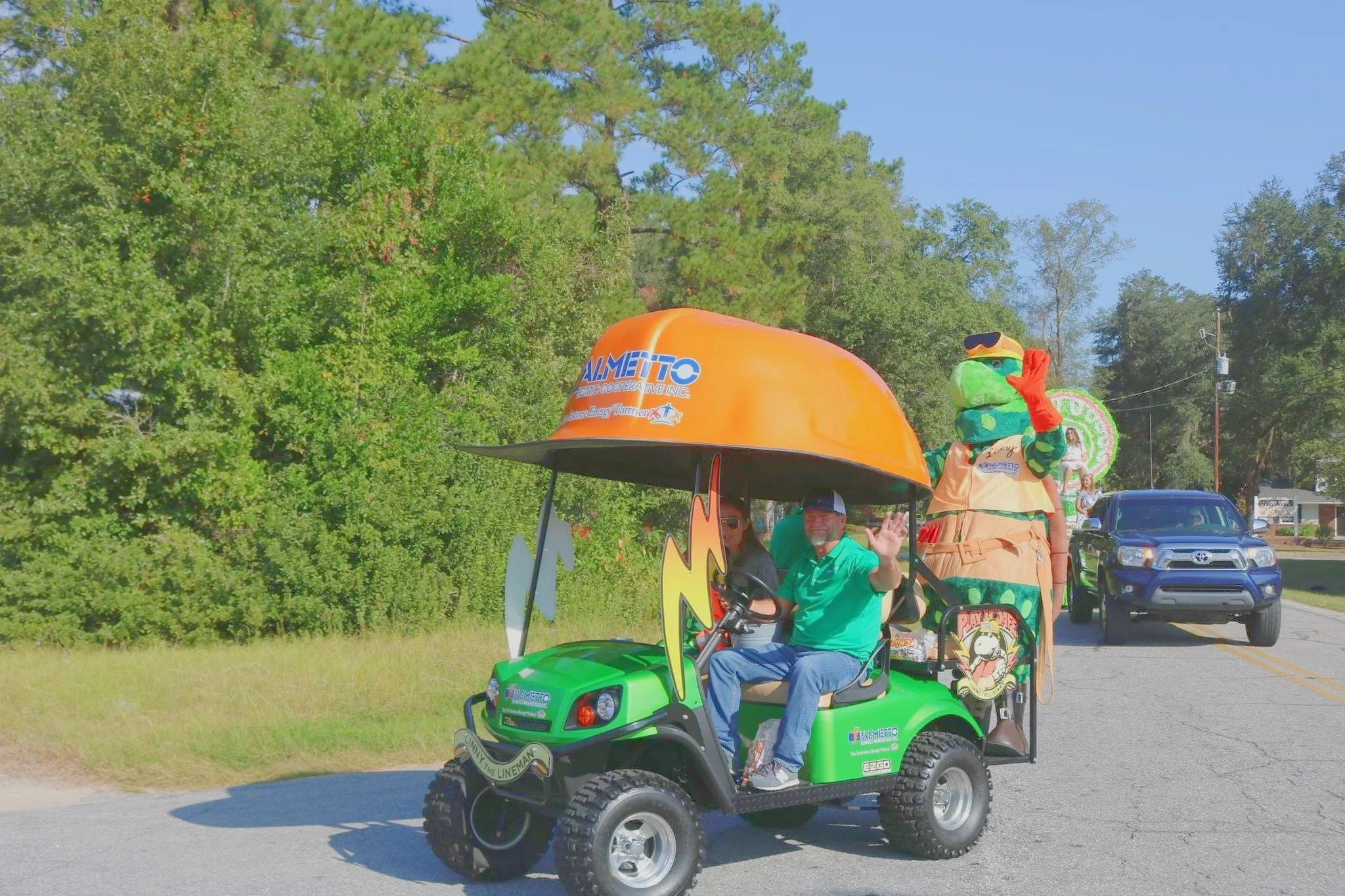 A small green golf cart with a person in a teal shirt waving, riding around during a parade. The golf cart has a mascot with sunglasses and a colorful costume standing behind the driver. There is a float in the background with a large orange umbrella and other parade participants. Trees and a blue sky are visible in the background.