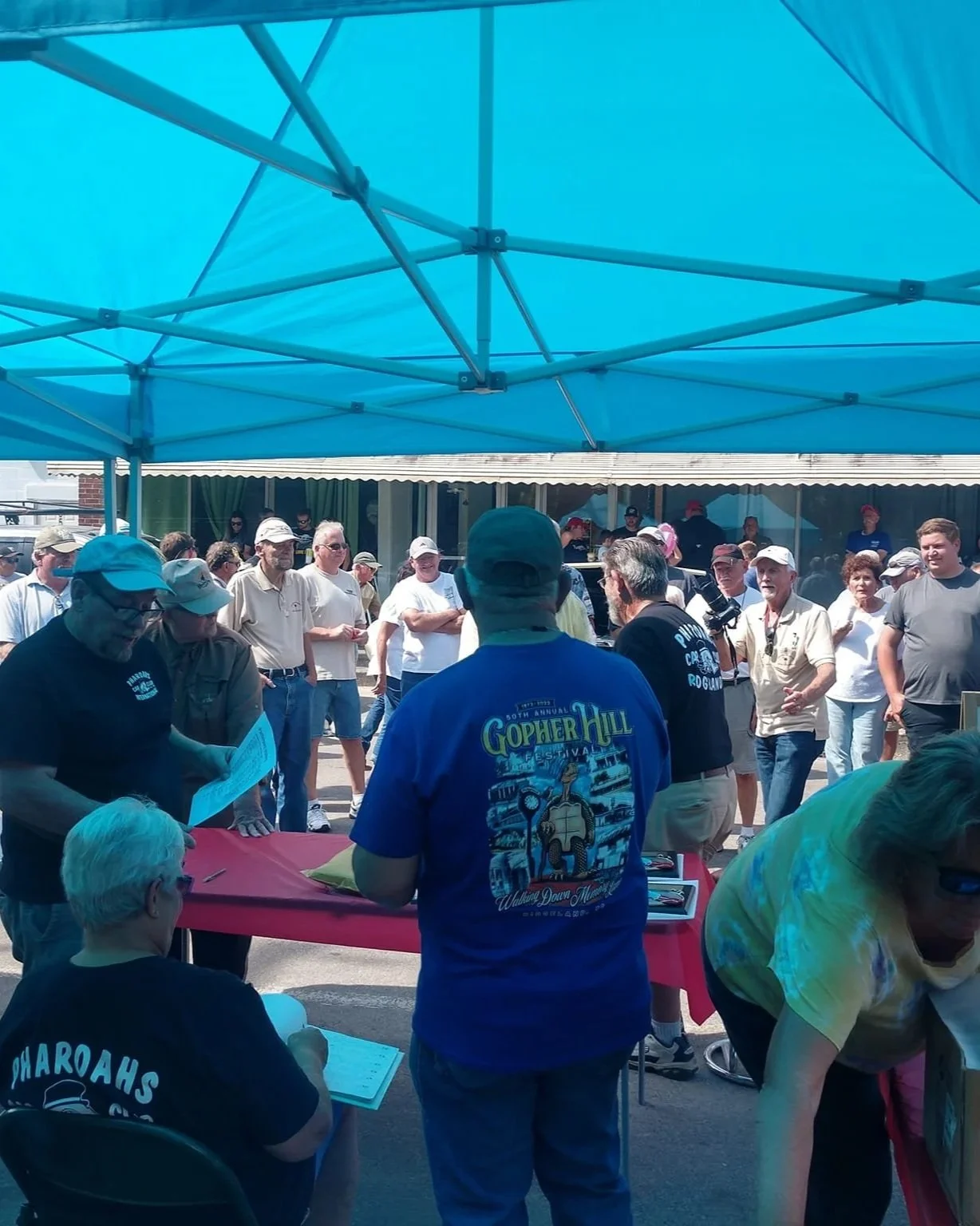 People gathered outdoors under a blue canopy, some wearing hats and casual clothing, at a festival or event with a red table, and a building with an awning in the background.