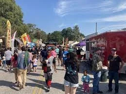 Crowd of people at an outdoor fair or festival with tents and flags under a blue sky.