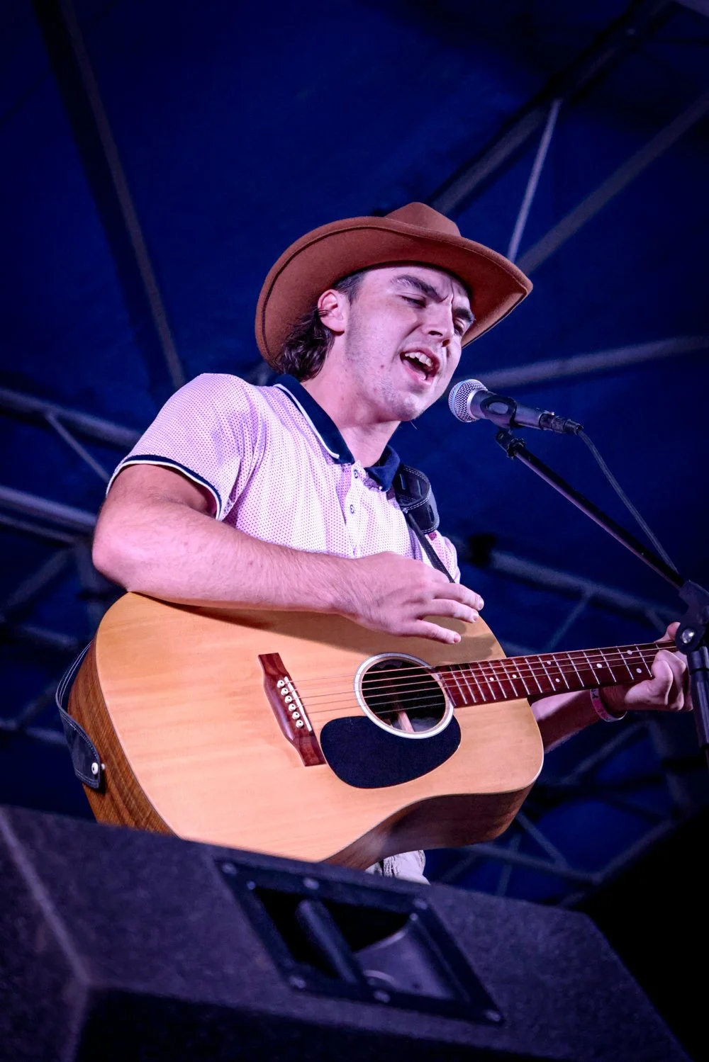 Young man performing on stage with a guitar, wearing a brown hat and a pink shirt, singing into a microphone under a blue canopy.