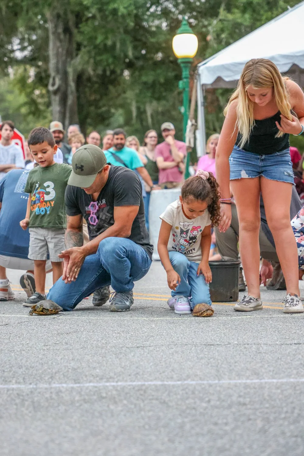 A man, woman, and children observing and interacting with turtles on a street during an outdoor event, with a crowd in the background.
